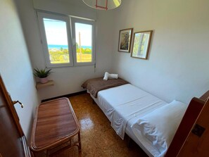 Quarto pequeno e arrumado com cama, cadeira, janela com vista e prateleira na parede com uma planta.