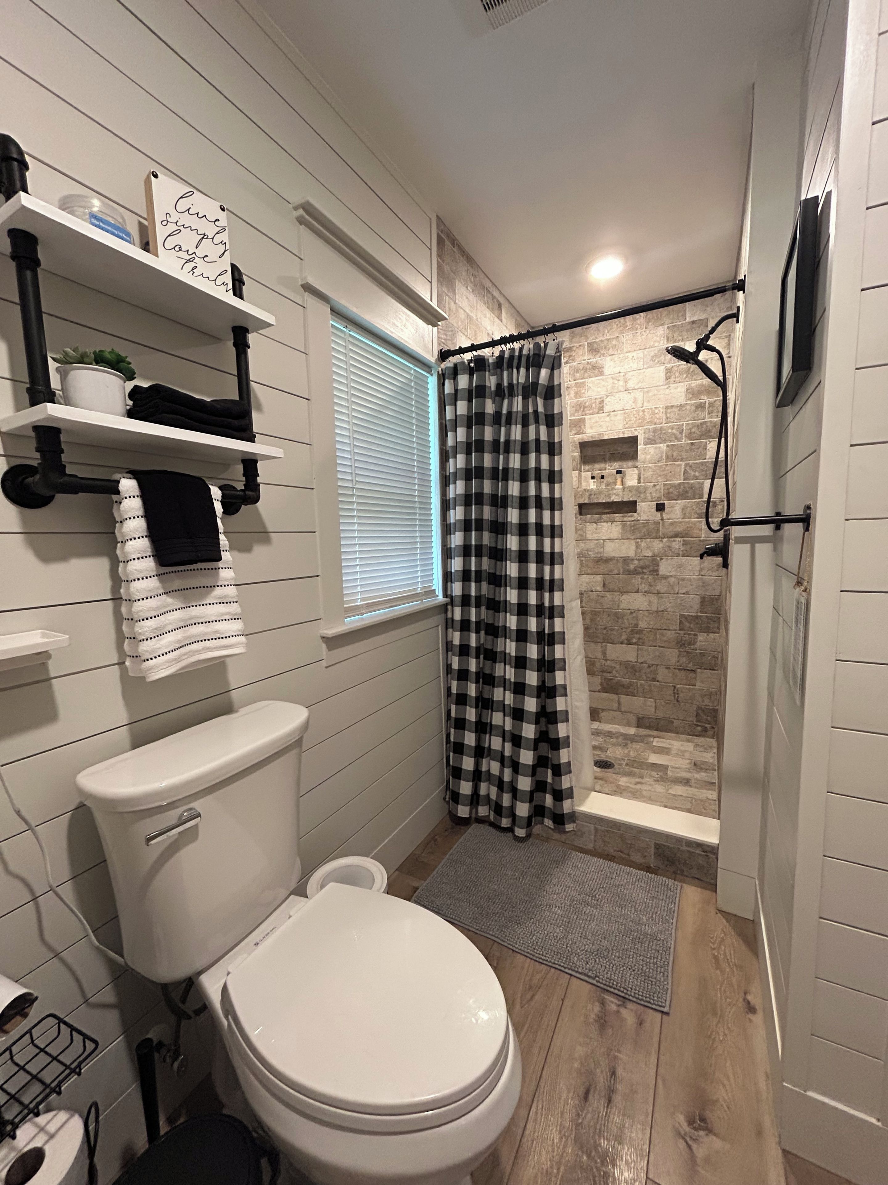 A bathroom with a white toilet, a shower with a checkered curtain, and a window with blinds.