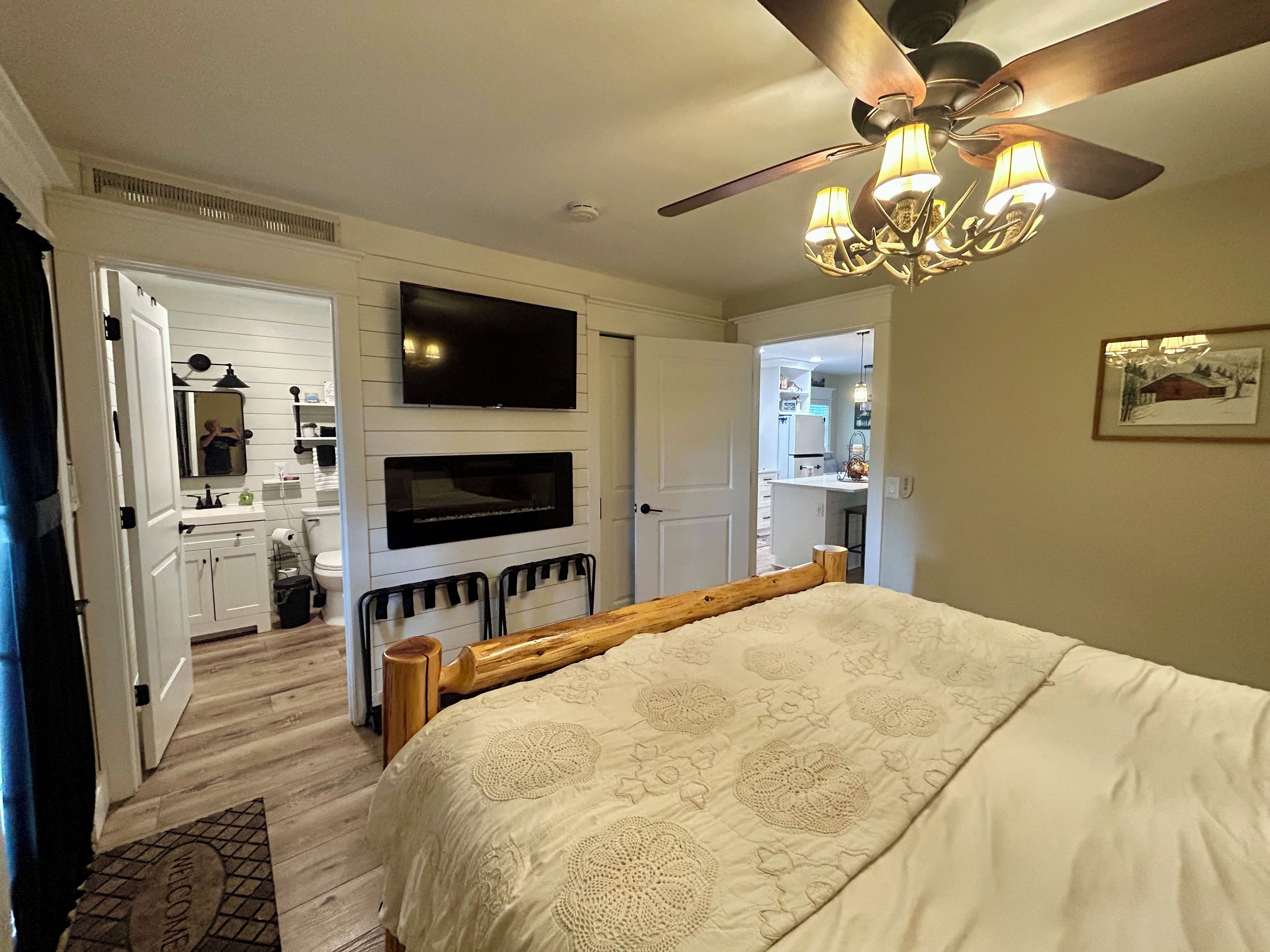A bedroom with a bed, a ceiling fan, a TV mounted above a fireplace, and a door leading to a bathroom.