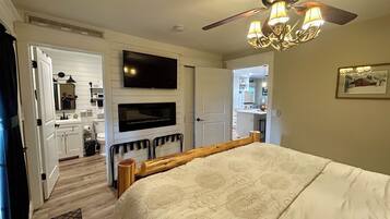 A bedroom with a bed, a ceiling fan, a TV mounted above a fireplace, and a door leading to a bathroom.
