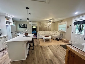 A modern kitchen with a central island, a dining area with a white table, and a living room with a sofa and a ceiling fan.