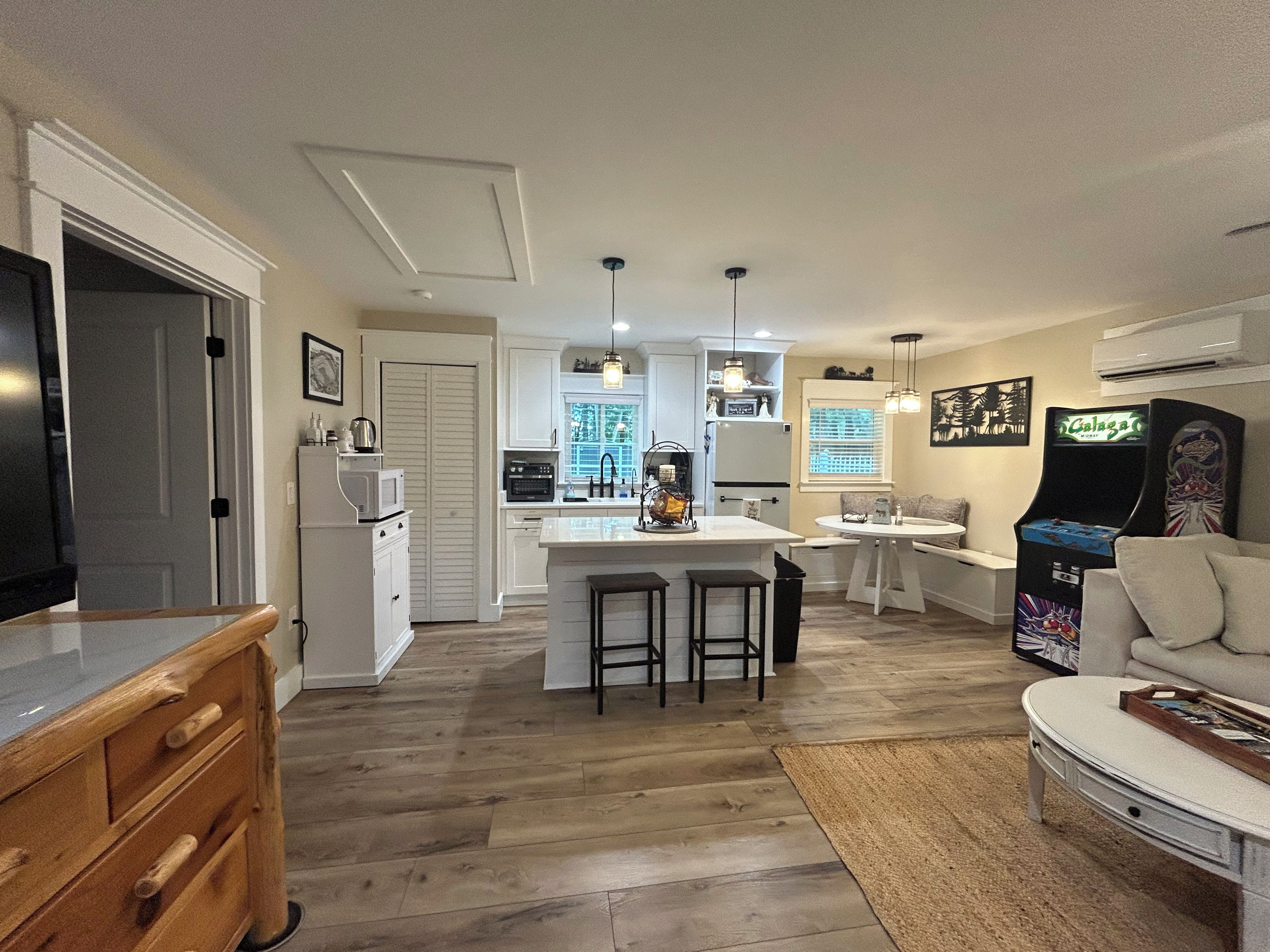 A modern kitchen with white cabinets, a central island with stools, a dining area with a table and chairs, and a retro arcade machine.