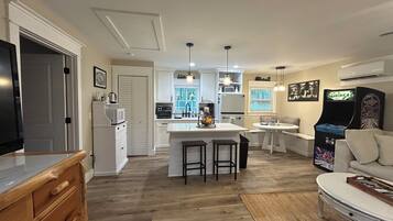 A modern kitchen with white cabinets, a central island with stools, a dining area with a table and chairs, and a retro arcade machine.