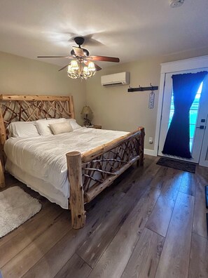 A bedroom with a wooden headboard, a ceiling fan, an air conditioning unit, and a door with a blue curtain. - Deer Haven Farmhouse-Cozy 1-Bedroom Cottage in Enchanting High Springs (High Springs)