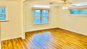 A room with hardwood flooring, white walls, a ceiling fan, and a large window. - Moss Side Cottage | 2BR • Huge Yard • Dogs Welcome (Baton Rouge)