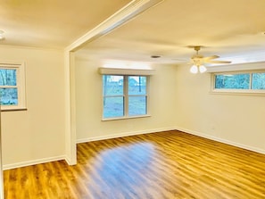 A room with hardwood flooring, white walls, a ceiling fan, and a large window. - Moss Side Cottage | 2BR • Huge Yard • Dogs Welcome (Baton Rouge)