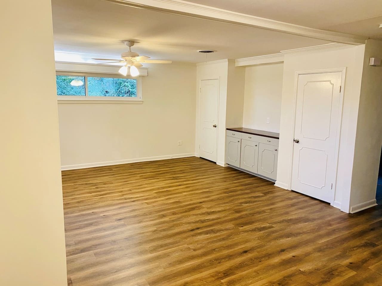 Quarto com piso de madeira, paredes brancas, ventilador de teto e janela com vista para árvores.
