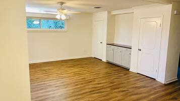 Quarto com piso de madeira, paredes brancas, ventilador de teto e janela com vista para árvores.