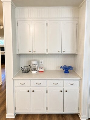 Cocina blanca con cafetera, una cesta y un objeto de vidrio azul sobre la encimera.