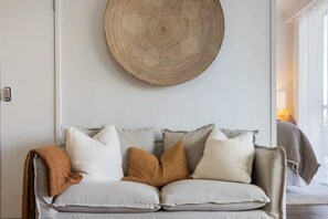 A neutral-colored sofa with multiple cushions against a plain wall, a round woven wall hanging above, and a glimpse of a window with curtains. - Ocea Coastal Luxe at Chevron (Surfers Paradise)