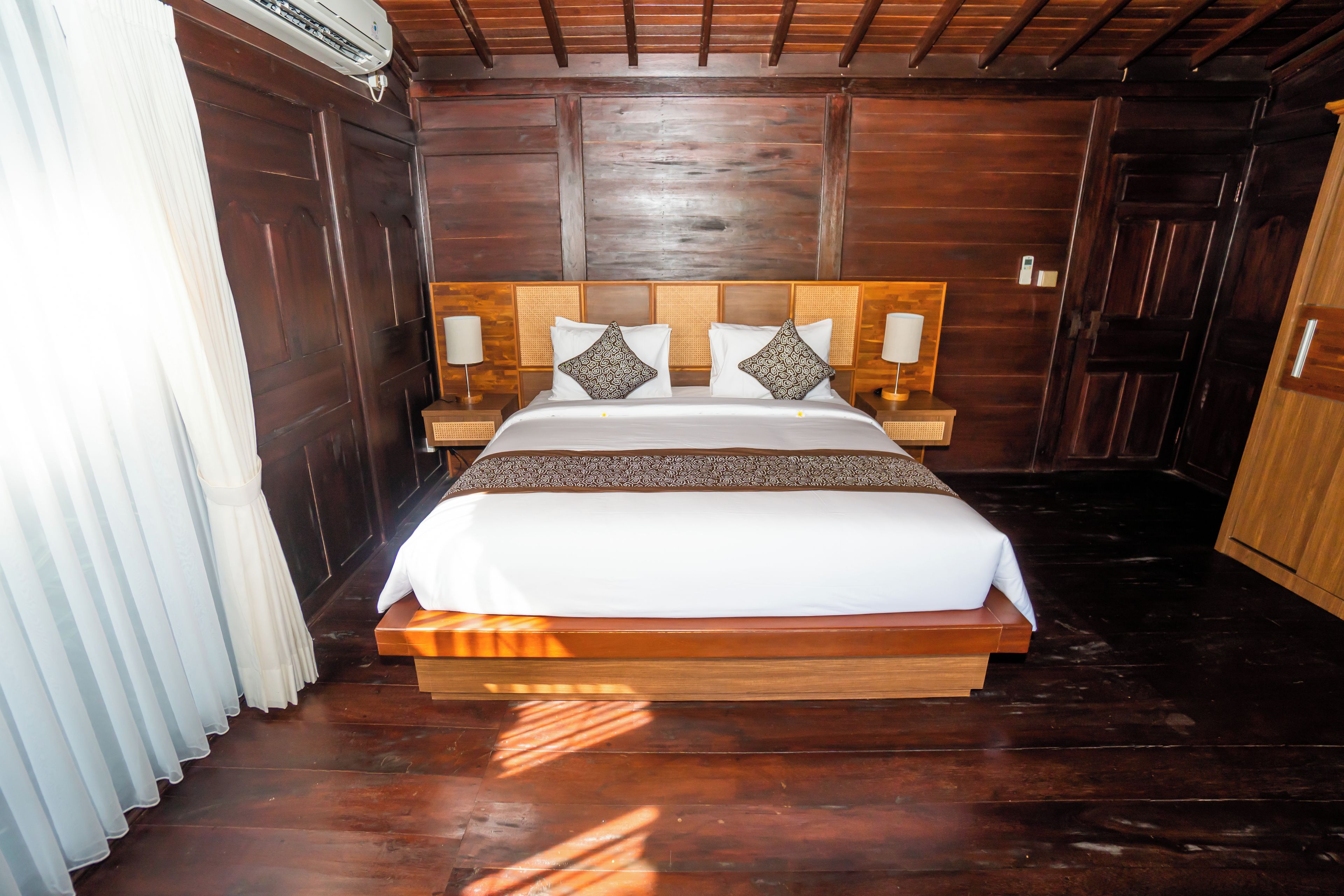 Uma cama de madeira com roupa de cama branca e almofadas estampadas, com mesas de cabeceira com abajures de cada lado, em um quarto com painéis de madeira e uma grande janela com cortinas.