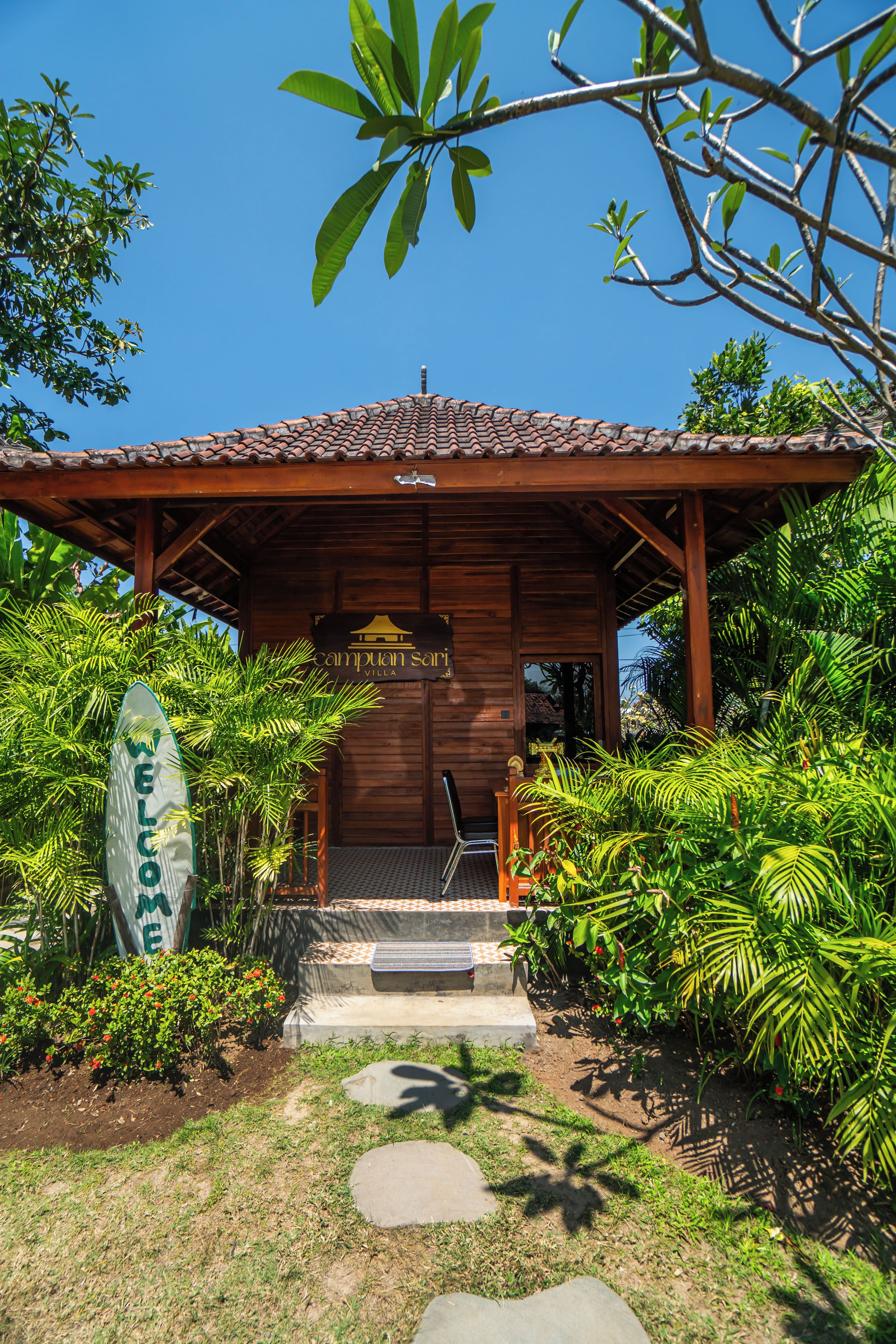 L’entrée d’une villa en bois, surmontée d’un toit en tuiles, est entourée d’une végétation luxuriante et s’ouvre sur un ciel d’un bleu limpide.