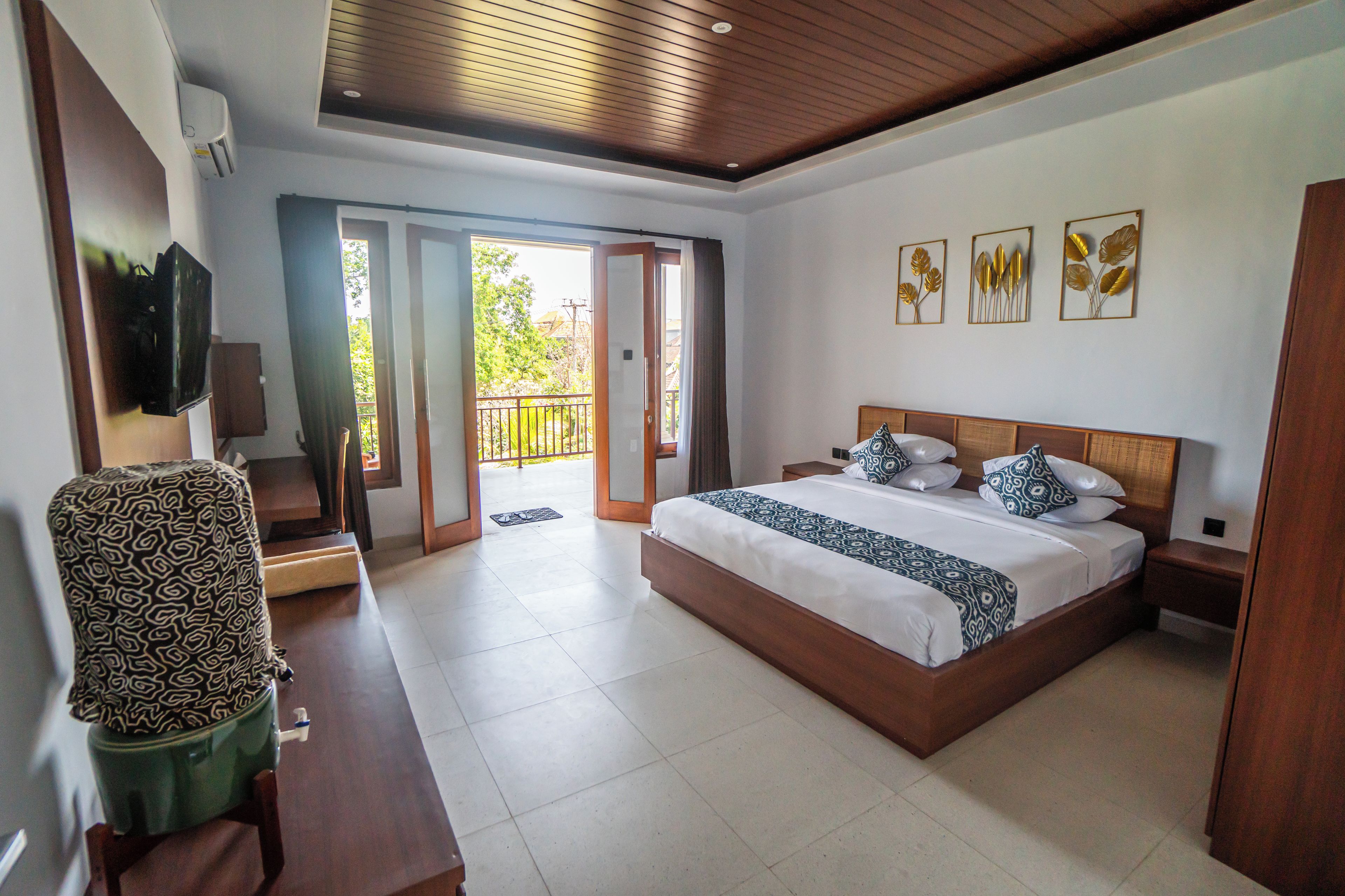 A bedroom with a large bed, a television, a desk, and a chair. There is a balcony with a view of greenery.