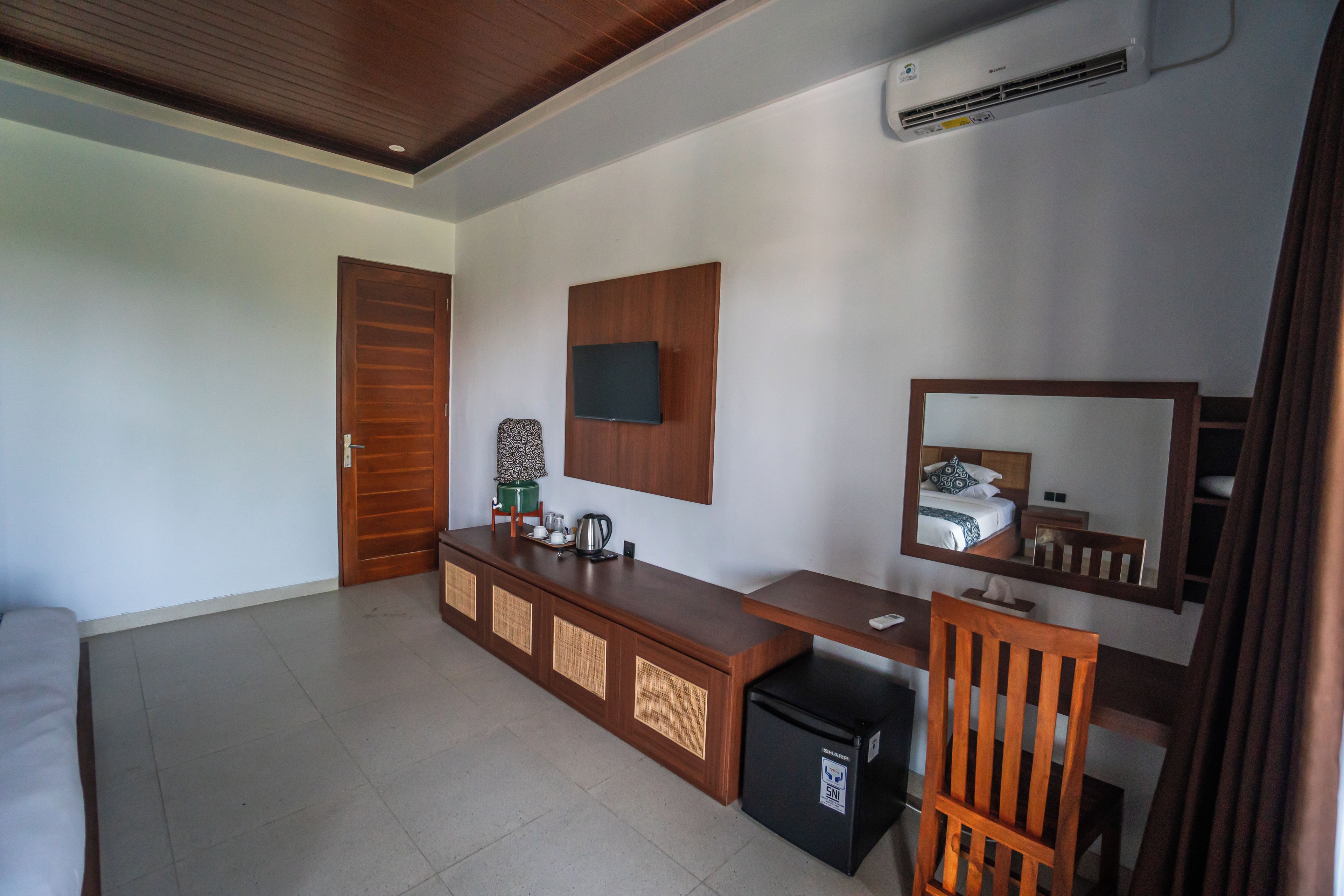 A hotel room with a wooden desk, a television mounted on the wall, a mirror, and a small refrigerator.