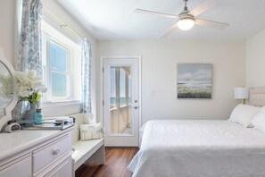 A bedroom with a bed, a bedside table, a chair, a window with curtains, a door, a ceiling fan, and a painting on the wall. - Ocean Reef 5-A1 | Oceanfront Condo (Emerald Isle)