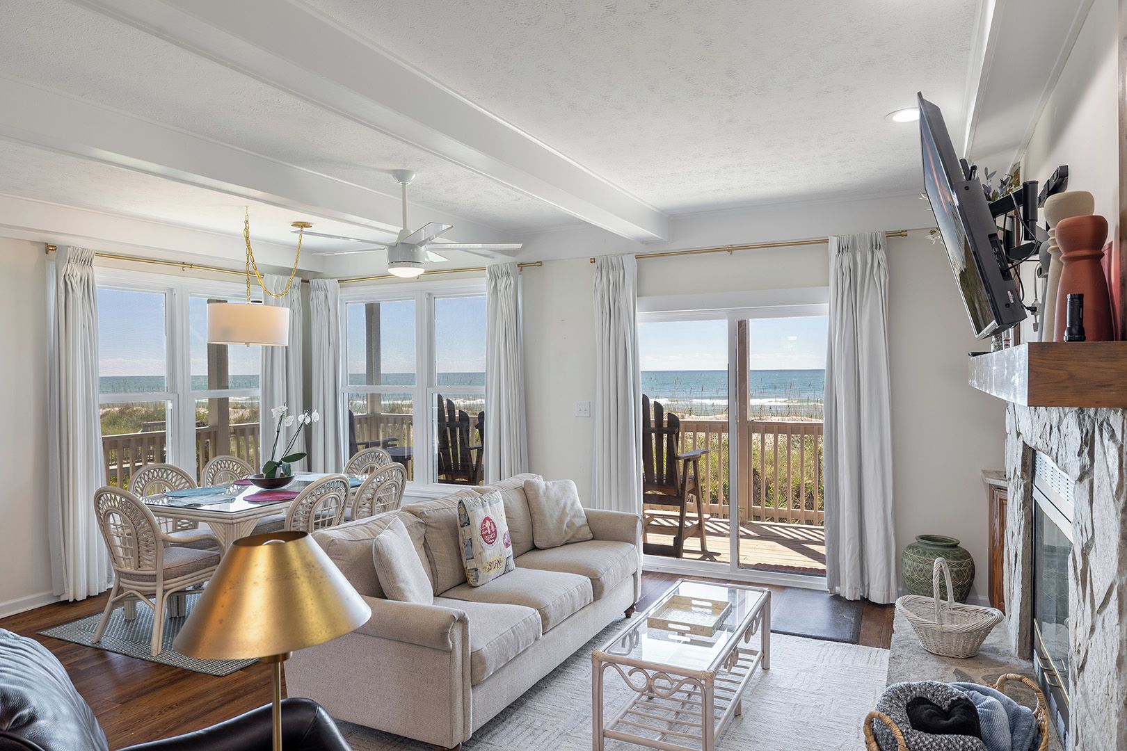 A spacious living area with a sofa, dining table, and chairs, a balcony with a view of the beach, and a television mounted on the wall.