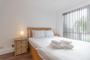 A bedroom with a wooden bed frame, white bedding, a nightstand with a lamp, and a window with vertical blinds. - Modern City Centre Apartment | Sleeps 4 | Parking - Pass the Keys (Chelmsford)