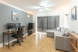 A modern living room with a desk, two monitors, a television, a grey sofa, and a large window with curtains. - Modern City Centre Apartment | Sleeps 4 | Parking - Pass the Keys (Chelmsford)