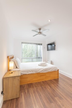 A bedroom with a wooden bed, a bedside table with a lamp, a ceiling fan, a window with blinds, and a wall-mounted TV. - Modern City Centre Apartment | Sleeps 4 | Parking - Pass the Keys (Chelmsford)