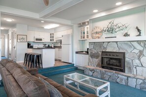 A modern living room with a stone fireplace, a kitchen with white cabinets, and a bar area with stools. - Pier Pointe 4B1 | Oceanfront 2 BR Condo (Emerald Isle)