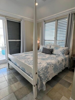 A four-poster bed with a patterned bedspread, a nightstand with a lamp, and a view of the outside through a window with blinds. - SlipAway TreasureCay (Treasure Cay)