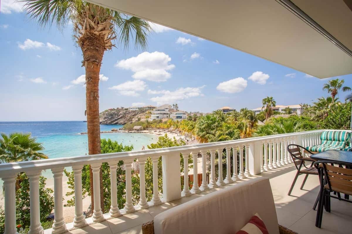 A balcony with a view of a beach and ocean, white balustrade, and outdoor furniture.