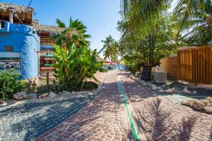 Un camí empedrat que condueix a una zona de platja, amb palmeres i un edifici amb teulada de palla.
