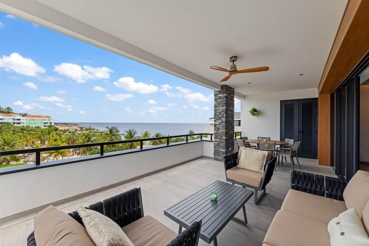 A balcony with a view of the ocean, a table, chairs, and a ceiling fan.