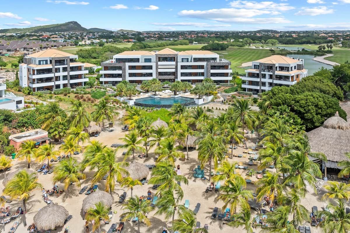 A resort with multiple buildings, a swimming pool, and a beach area with palm trees and sun loungers.