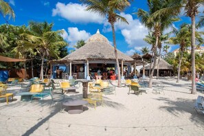 A beachside area with thatched-roof structures, outdoor seating, and palm trees. - NEW: Beachfront Blue Bay Resort The Shore 6 (Sint Michiel)