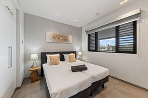 A modern bedroom with a large bed, two bedside tables, and a window with blinds. - NEW: Beachfront Blue Bay Resort The Shore 6 (Sint Michiel)