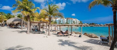 Een strand met palmbomen, een hut met rieten dak en een gebouw met balkons.
