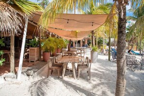 Un área de estar al aire libre con mesas y sillas de madera, protegida por un toldo grande, rodeada de plantas en macetas y palmeras.