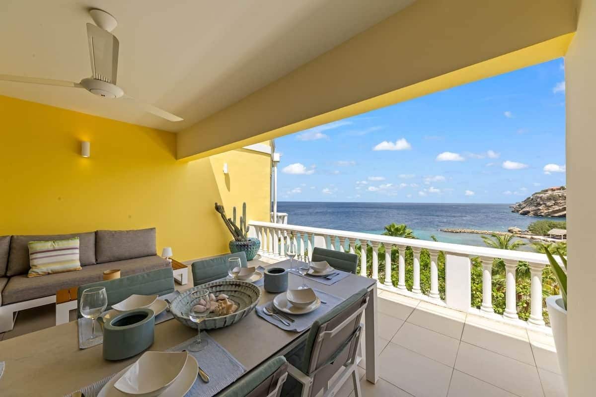 A balcony with a dining table set for two, a sofa, and a view of the sea.