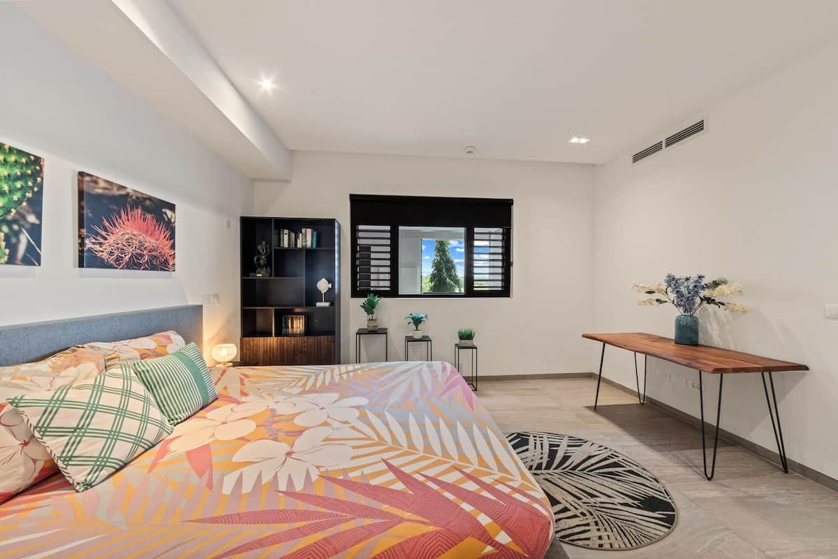 A bedroom with a bed, a nightstand, a window with blinds, and a small table with a vase of flowers.