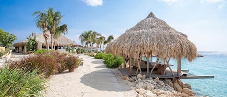 A beachside resort with thatched-roof structures, a paved walkway, and a view of the sea.