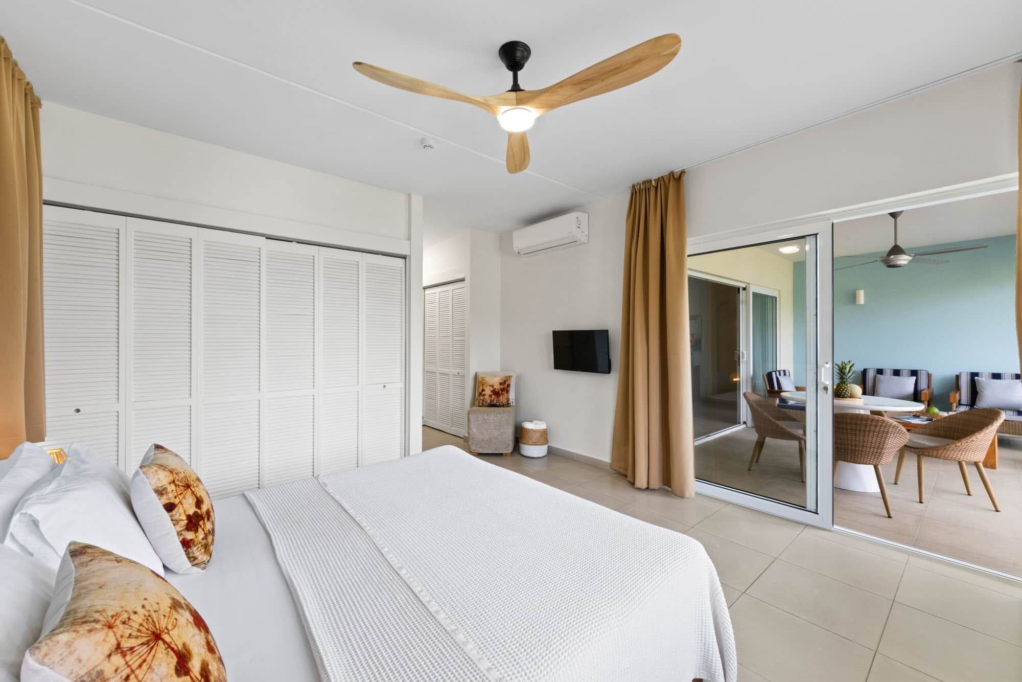 A hotel room with a bed, a ceiling fan, a TV, and a sliding glass door leading to a balcony with seating.