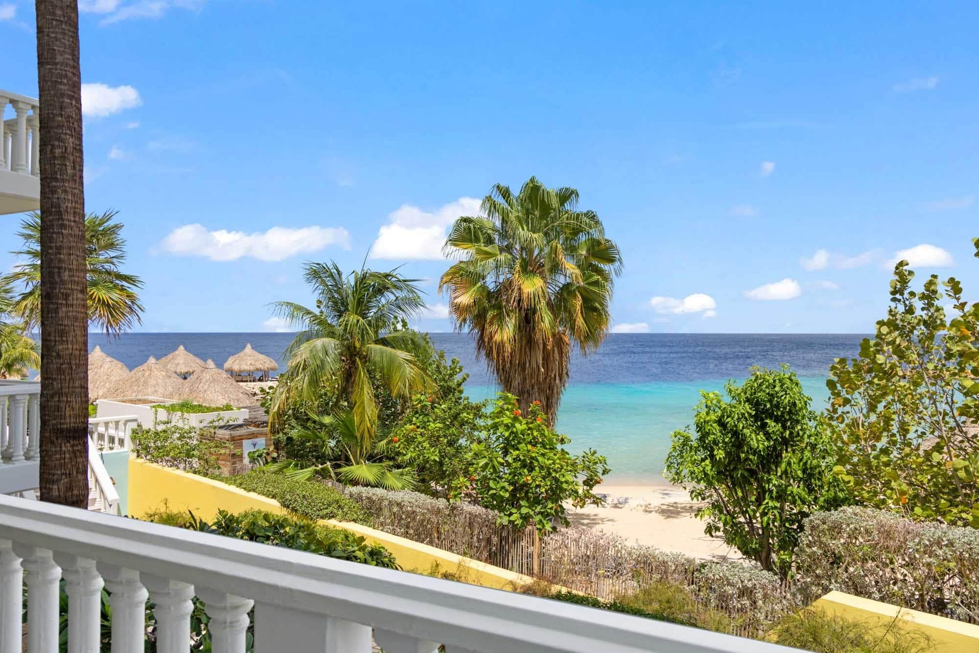 A beachfront property with a balcony overlooking the ocean, palm trees, and a thatched-roof structure.