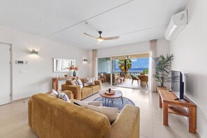 Ein modernes Wohnzimmer mit Sofa, Couchtisch und Fernseher. Ein Balkon bietet Blick aufs Meer.