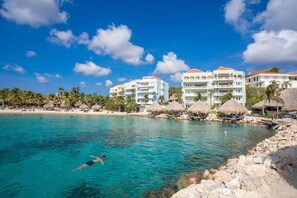 Ein Resort mit klarem türkisblauem Wasser, einer schwimmenden Person und Strandgebäuden mit Strohdächern.