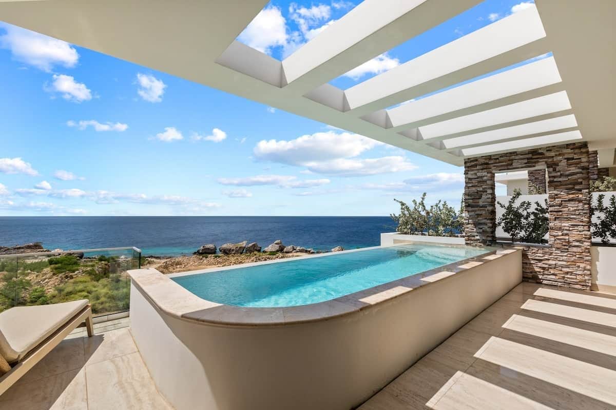 A pool with a view of the ocean, a stone wall, and a pergola structure.