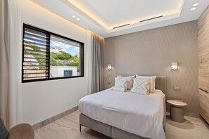 A modern bedroom with a large bed, a bedside table, a window with blinds, and a view of greenery outside. - NEW: The Ridge, Penthouse on The Blue Bay Resort (Sint Michiel)
