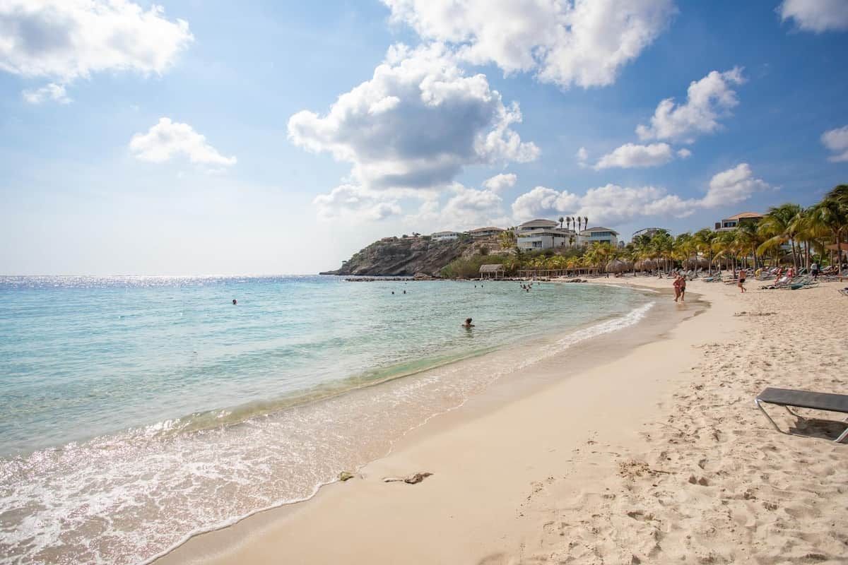 Una playa con agua cristalina, una orilla arenosa y un edificio de varios pisos sobre una colina.