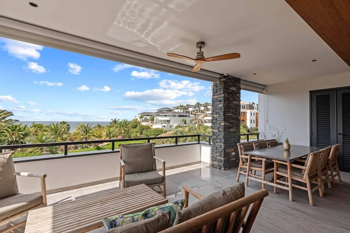 A balcony with a table and chairs, a ceiling fan, and a view of the ocean and buildings.