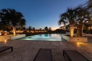 A pool area with lounge chairs and palm trees, surrounded by a building with warm lighting. - NEW: The Shore Beachfront Blue Bay Resort 3 BDR (Sint Michiel)