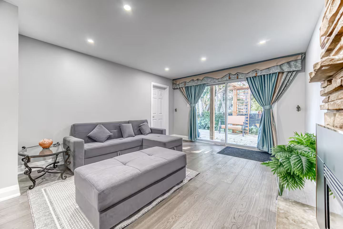Una sala de estar moderna con un sofá seccional gris, una mesa de centro de vidrio y un otoman gris. Hay un amplio ventanal corredero que da a un balcón con vistas al exterior.