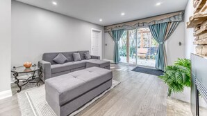 Uma sala de estar moderna com um sofá sectional cinza, uma mesa de centro de vidro e um banquinho cinza. Há uma grande porta de vidro deslizante que dá para uma varanda com vista para o exterior.