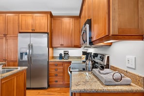 A kitchen with wooden cabinets, a stainless steel refrigerator, a coffee maker, a kettle, and granite countertops. - Orange Crush - Private Pool Home Near Beach - By Island Real Estate (Holmes Beach)