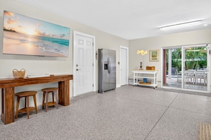 A kitchen with a refrigerator, a dining table with stools, a painting, and a sliding glass door leading to an outdoor area. - Orange Crush - Private Pool Home Near Beach - By Island Real Estate (Holmes Beach)
