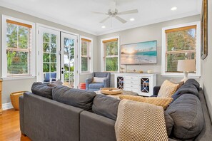 Ein Wohnzimmer mit einem grauen Ecksofa, einem gemusterten Sessel, einer weißen Kommode und einem großen Fenster mit Jalousien.